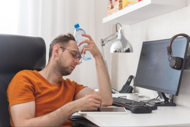 Man in his home office holding a water bottle to his forehead