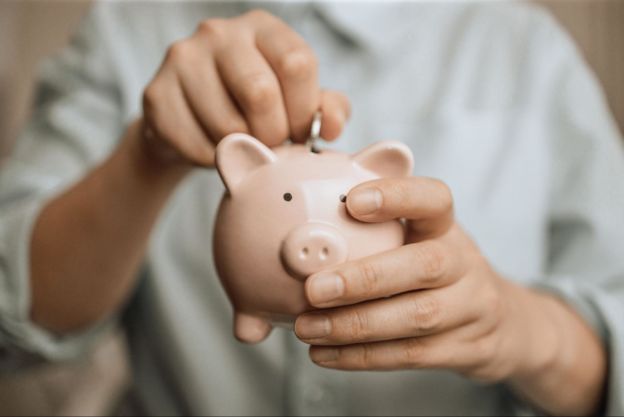 A man holding a light pink piggy bank & putting a coin in it.